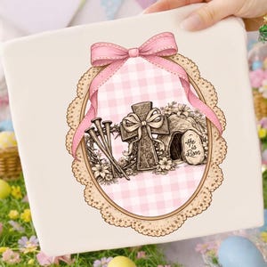 May include: A cream gift box with an Easter illustration. The design features a cross, nails, and a tomb with the text "He is Risen." A pink bow and ribbon frame the image, set against a pink gingham background within a decorative oval.