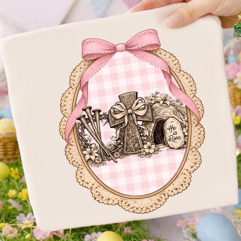 May include: A cream gift box with an Easter illustration. The design features a cross, nails, and a tomb with the text "He is Risen." A pink bow and ribbon frame the image, set against a pink gingham background within a decorative oval.