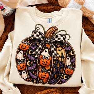 Op de afbeelding: Een crèmekleurig sweatshirt met ronde hals en een grote afbeelding van een pompoen versierd met een zwart-witte ruitjesstrik. Het pompoenontwerp bevat illustraties van lantaarns, witte spoken, paarse vleermuizen, gele maansikkels en een donker spookhuis.