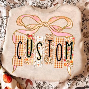 May include: A beige sweatshirt with the word "CUSTOM" in black block letters. The letters are designed with floral, plaid, and patterned fabric. A large pink bow with gold glitter accents is above the word.