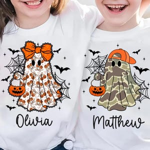 以下が含まれることがあります： ハロウィーンのゴーストデザインの白いTシャツ。1つのゴーストはオレンジ色のリボンとカボチャの模様があり、もう1つは迷彩模様とオレンジ色のキャップを着用しています。どちらもジャック・オー・ランタンのバッグを持っています。黒いコウモリとクモの巣がシャツを飾っています。オリビアとマシューの名前が印刷されています。