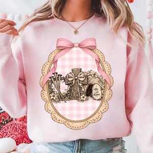 May include: A pale pink sweatshirt with an Easter graphic. The design features a cross, nails, and a tomb with the text "He is Risen." The graphic is framed by a pink gingham oval and a pink bow.