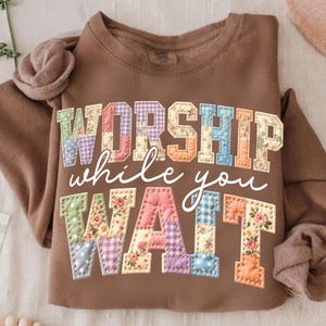 Peut inclure: Un sweat-shirt marron avec l'inscription "WORSHIP while you WAIT" dans un style patchwork. Les lettres sont faites de tissus floraux et à motifs, avec un aspect cousu. Le sweat-shirt a des manches longues et un col rond.