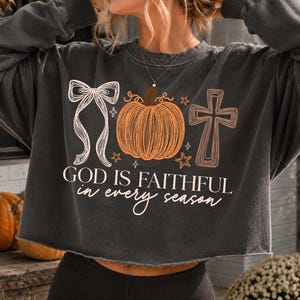 May include: A charcoal gray cropped sweatshirt featuring a graphic print with a white ribbon bow, an orange pumpkin, and a brown cross. The text reads "GOD IS FAITHFUL in every season" in white and light orange lettering with small star accents.