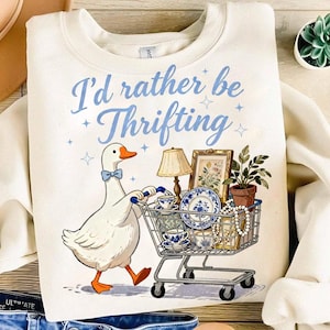 May include: Cream-colored sweatshirt with the text "I'd rather be Thrifting" in blue script. A cartoon goose with a blue bow tie pushes a shopping cart filled with vintage items, including a lamp, framed art, and teacups.