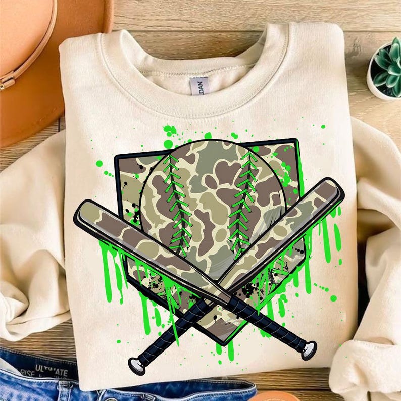 May include: A beige sweatshirt featuring a green, white and brown camouflage baseball and two crossed bats on a white home plate. The design is surrounded by green paint splatters.