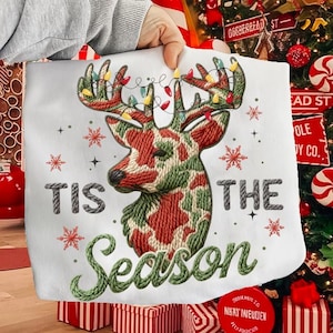 May include: A white Christmas pillow featuring an embroidered reindeer design with red and green camouflage-style patterns. The reindeer's antlers are adorned with string lights. The text "TIS THE SEASON" is embroidered in green and gray.