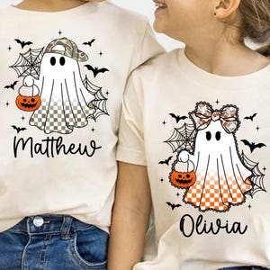 Puede incluir: Dos camisetas de Halloween color crema. Una presenta un fantasma con gorra de camuflaje, sosteniendo una calabaza, con el nombre "Matthew" debajo. La otra tiene un fantasma con un lazo, sosteniendo una calabaza, con el nombre "Olivia".