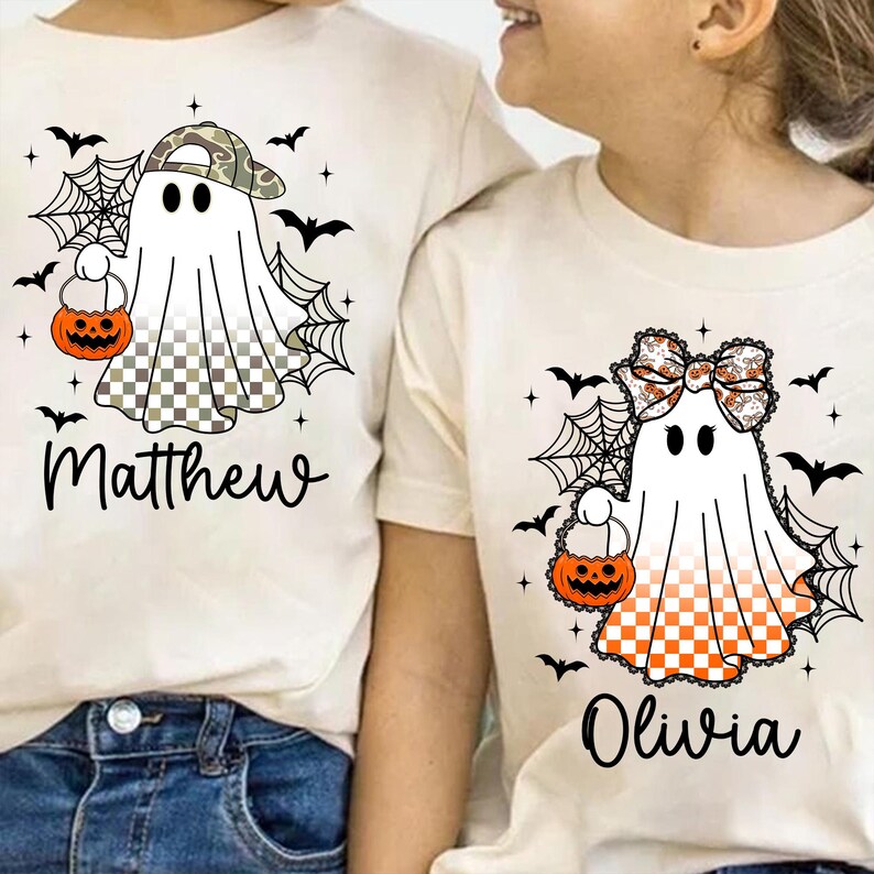 Puede incluir: Dos camisetas de Halloween color crema. Una presenta un fantasma con gorra de camuflaje, sosteniendo una calabaza, con el nombre "Matthew" debajo. La otra tiene un fantasma con un lazo, sosteniendo una calabaza, con el nombre "Olivia".