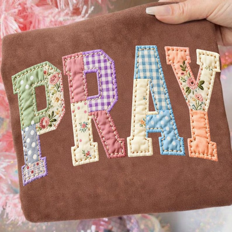 May include: Brown fabric with the word "PRAY" appliqued in colorful patchwork letters. The letters feature floral, polka dot, and gingham patterns in shades of pink, blue, green, and purple. The letters are outlined with a cream-colored stitching.