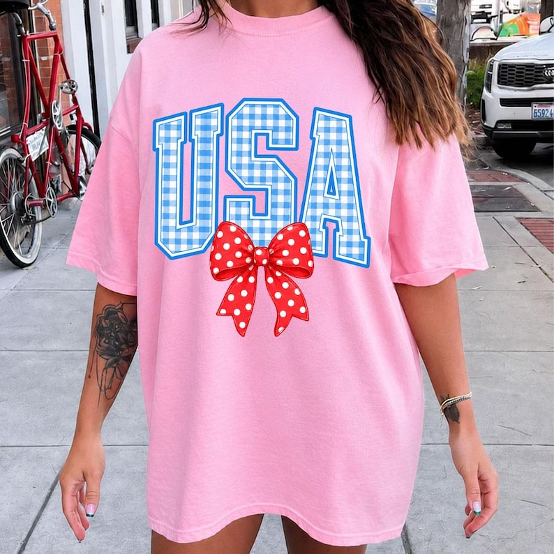 May include: A pink t-shirt with the word "USA" in blue gingham letters. A red bow with white polka dots is centered below the text. The shirt has short sleeves and a relaxed fit.
