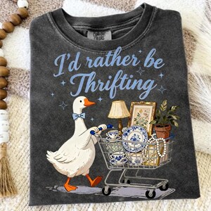 May include: Dark gray t-shirt with the phrase "I'd rather be Thrifting" in light blue. A cartoon goose with a blue bow tie pushes a shopping cart filled with vintage items, including teacups, plates, and a framed picture.