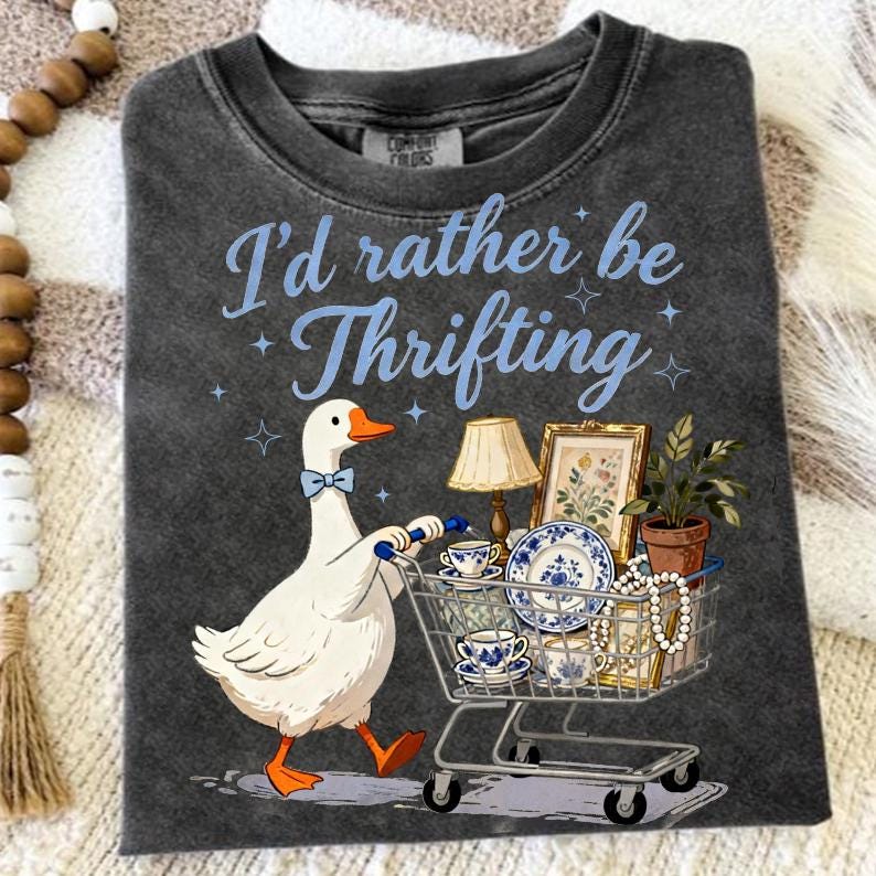 May include: Dark gray t-shirt with the phrase "I'd rather be Thrifting" in light blue. A cartoon goose with a blue bow tie pushes a shopping cart filled with vintage items, including teacups, plates, and a framed picture.
