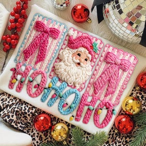 Peut inclure: Textile blanc sur le thème de Noël avec des broderies. Il présente un visage de Père Noël, des nœuds roses et le texte "HO HO HO" en bleu et rose, orné de guirlandes lumineuses. Des boules rouges et dorées entourent le textile.