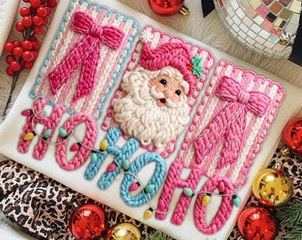Puede incluir: Textil blanco con bordados navideños. Presenta una cara de Papá Noel, lazos rosas y el texto "HO HO HO" en azul y rosa, adornado con luces de cadena. Adornos rojos y dorados rodean el textil.
