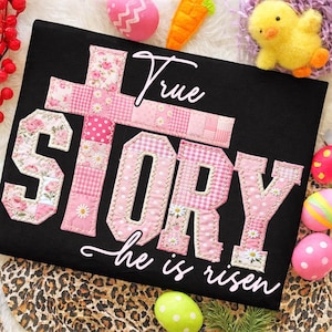 May include: Black t-shirt with the words "True Story" in a patchwork design, featuring a cross and letters in pink floral and gingham patterns. The phrase "he is risen" is written below. Easter-themed decorations surround the shirt.
