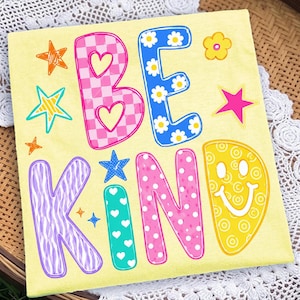 Op de afbeelding: Een lichtgeel T-shirt met de tekst BE KIND in grote, kleurrijke, gedessineerde letters. De B is roze geblokt met hartjes, de E is blauw met madeliefjes, de K is paars met zebrastrepen, de I is groenblauw met witte hartjes, de N is roze met witte stippen en de D is een geel smileygezicht.