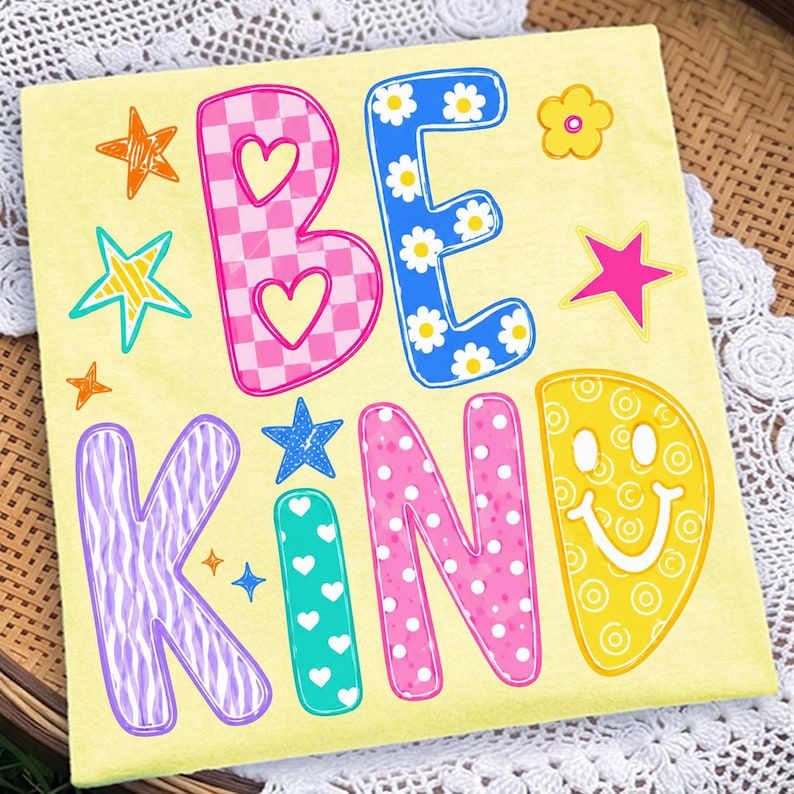 Op de afbeelding: Een lichtgeel T-shirt met de tekst BE KIND in grote, kleurrijke, gedessineerde letters. De B is roze geblokt met hartjes, de E is blauw met madeliefjes, de K is paars met zebrastrepen, de I is groenblauw met witte hartjes, de N is roze met witte stippen en de D is een geel smileygezicht.