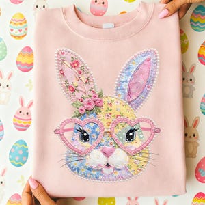 May include: A light pink sweatshirt featuring a patchwork bunny face with heart-shaped glasses. The bunny has floral patterned ears and a nose, with a small flower crown. The sweatshirt is folded and held by hands.