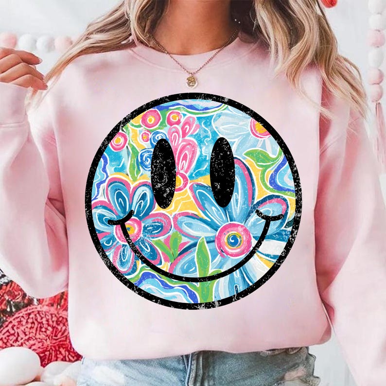 May include: A light pink sweatshirt featuring a large smiley face design. The smiley face is filled with a colorful floral pattern in shades of blue, pink, yellow, and green, outlined in black.