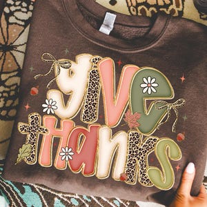 May include: A brown sweatshirt with the words "GIVE THANKS" in a decorative font. The letters are in various colors, including pink, green, and white, with leopard print accents. The design includes floral and leaf elements.