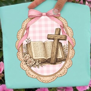 May include: A light blue decorative pillow featuring a pink and white gingham pattern, a wooden cross, and an open book. The design includes a pink bow and a floral border, creating a religious theme.