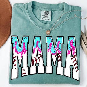 Peut inclure: Un t-shirt bleu turquoise avec le mot "MAMA" en lettres blanches avec un design sur le thème du baseball rose et bleu.