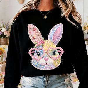 May include: Black sweatshirt featuring a colorful bunny face with heart-shaped glasses. The bunny design includes floral patterns in pink, yellow, and blue. The sweatshirt is a casual style, suitable for spring or Easter.