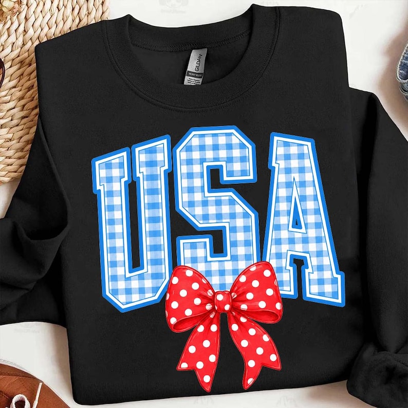 May include: Black sweatshirt with the word "USA" in blue and white gingham print. A red bow with white polka dots is at the bottom of the letters. The sweatshirt is a Gildan brand.