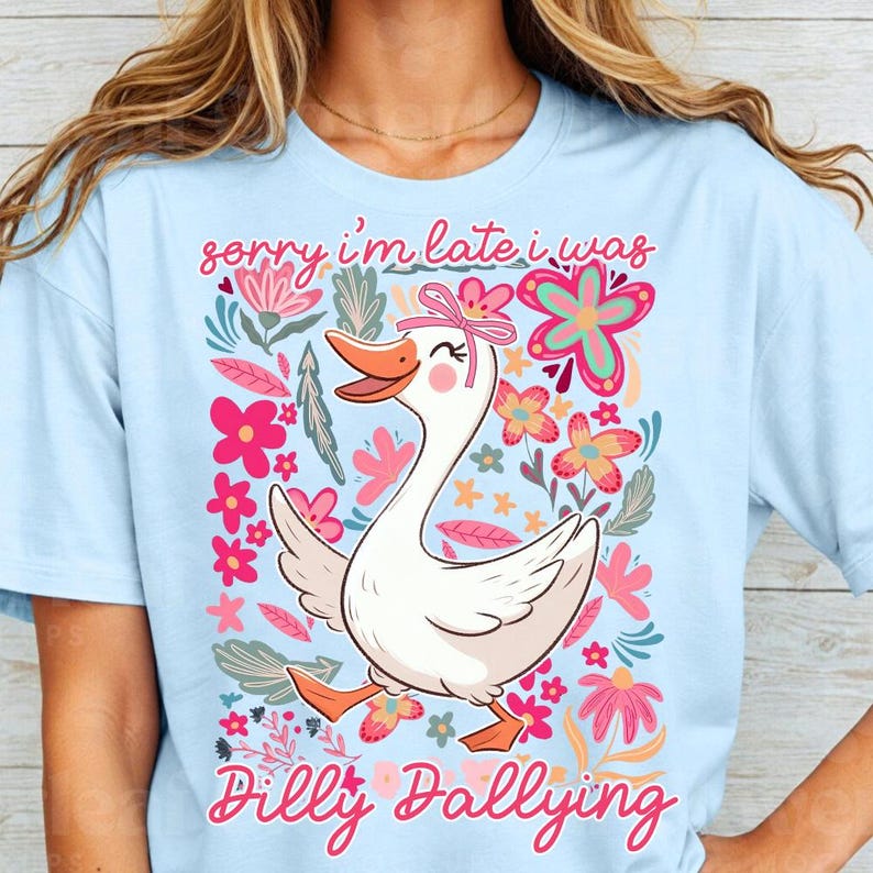 K&ouml;nnte beinhalten: Hellblaues T-Shirt mit einem Cartoon-Gans-Design, die eine rosa Schleife tr&auml;gt und von Blumen umgeben ist. Der Text lautet "Sorry I'm late I was Dilly Dallying" in rosa Schrift. Ein verspieltes und farbenfrohes Design.