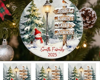 Peut inclure: Ornement de Noël rond avec une scène hivernale. Il présente un bonhomme de neige, un lampadaire et un poteau indicateur avec des noms. L'ornement porte l'inscription "Smith Family 2025". Un oiseau rouge est perché sur le poteau.