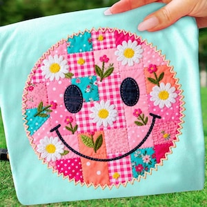 Könnte beinhalten: Ein hellblaues Stoffstück mit einem Patchwork-Smiley-Gesicht-Design. Das Smiley-Gesicht besteht aus verschiedenen gemusterten Quadraten in Rosa, Türkis und Orange, mit Gänseblümchen- und Blumenstickereien. Die Kanten sind mit einem dekorativen Zickzackstich versehen.