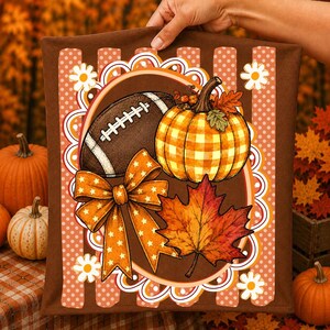 May include: A brown decorative cushion cover featuring a central oval frame with a football, a yellow and orange chequered pumpkin, a large orange bow with white stars, and a maple leaf, set against a background of vertical stripes and dots.