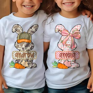May include: Two white t-shirts with Easter bunny graphics. One bunny has a camouflage pattern with a green hat and the name "andrew". The other bunny has a pink floral pattern with a bow and the name "amelia". Each shirt has a carrot graphic.