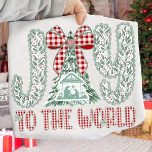 May include: White sweatshirt with a Christmas-themed design. Features the words "JOY TO THE WORLD" in a red and white plaid pattern. The design includes a Christmas tree, a bow, and a nativity scene.