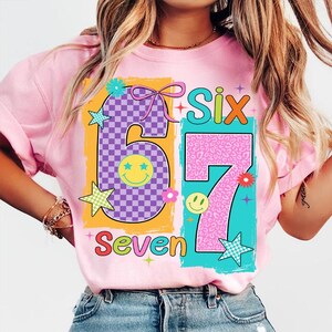 May include: Pink t-shirt with a colourful graphic design featuring the numbers "6" and "7" in a playful font. The number "6" has a checkered pattern, and the number "7" has a leopard print. The words "Six" and "Seven" are also included.
