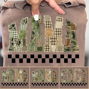 Op de afbeelding: Een bruine sweatshirt met het woord "MAMA" in een patchwork-ontwerp. De letters zijn gemaakt van verschillende camouflage- en ruitstoffen. Onder de letters bevindt zich een zwart-wit geblokt patroon.