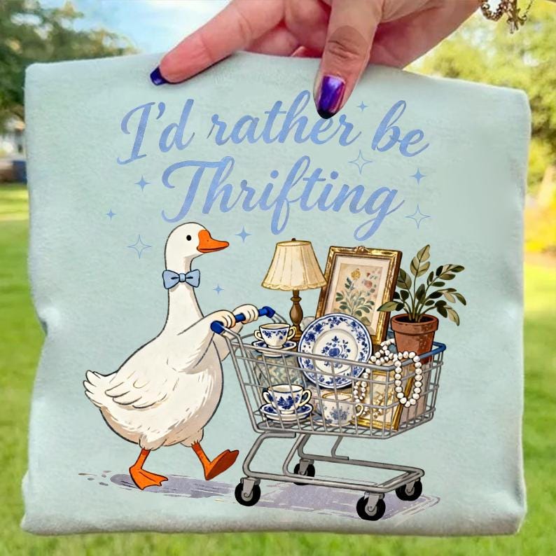 May include: Light blue t-shirt with the text "I'd rather be Thrifting." A cartoon goose with a blue bow tie pushes a shopping cart filled with vintage items, including a lamp, framed art, and blue and white china.