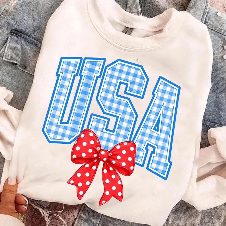May include: White sweatshirt with the word "USA" in blue gingham letters. A red bow with white polka dots is centered below the text. The sweatshirt is displayed on a denim jacket.