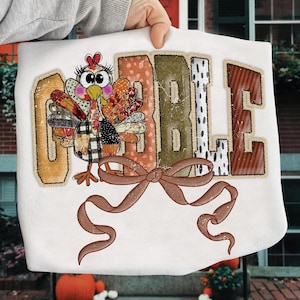 Könnte beinhalten: Weißes Sweatshirt mit einer bunten Truthahn-Grafik und dem Wort "GOBBLE" im Used-Patchwork-Stil. Der Truthahn hat einen karierten Körper und eine braune Schleife. Ein festliches Design für Thanksgiving.