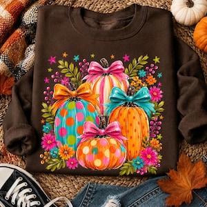 May include: A dark brown long-sleeve sweatshirt featuring a central graphic of four pumpkins in orange, teal, and white, each decorated with colorful polka dots and topped with a large, vibrant bow in shades of pink, orange, or teal.