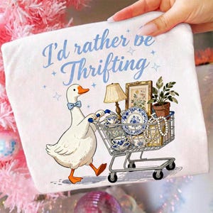 May include: White graphic tee featuring a cartoon goose pushing a shopping cart filled with vintage items. The text "I'd rather be Thrifting" is written in blue script. The goose wears a blue bow tie.