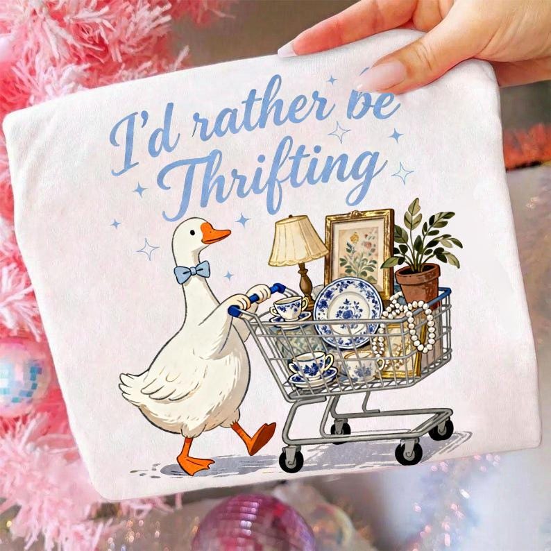 May include: White graphic tee featuring a cartoon goose pushing a shopping cart filled with vintage items. The text "I'd rather be Thrifting" is written in blue script. The goose wears a blue bow tie.