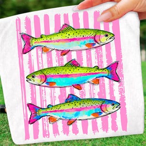 May include: White towel featuring three colorful rainbow trout illustrations. The fish have green backs, pink and blue stripes, and bright pink fins. The background has vertical pink stripes.