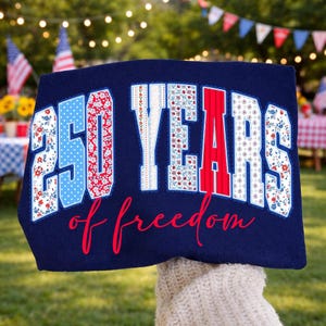 Pode incluir: Um tecido azul marinho com o texto "250 YEARS of freedom". Os números e letras estão em padrões vermelhos, brancos e azuis, com detalhes florais e de bolinhas. O fundo é uma cena desfocada ao ar livre.