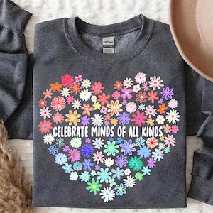 May include: A dark gray sweatshirt featuring a heart-shaped floral design in various colors. The heart is composed of numerous flowers, with the words "CELEBRATE MINDS OF ALL KINDS" printed in white within the heart. The sweatshirt has long sleeves.