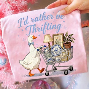 May include: Pink t-shirt with the text "I'd rather be Thrifting" and a cartoon goose pushing a shopping cart filled with vintage items. The cart includes a lamp, teacups, and a framed picture.