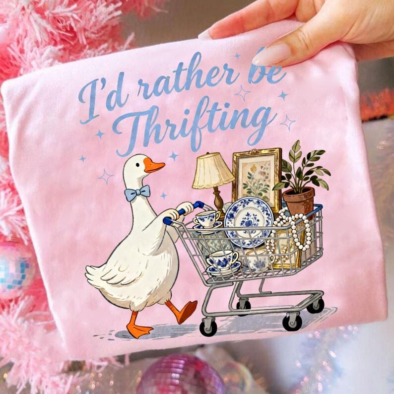 May include: Pink t-shirt with the text "I'd rather be Thrifting" and a cartoon goose pushing a shopping cart filled with vintage items. The cart includes a lamp, teacups, and a framed picture.