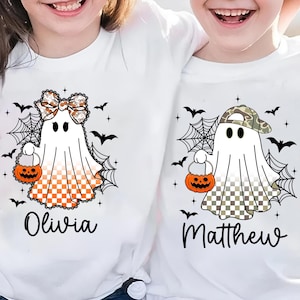 Puede incluir: Camisetas blancas con dise&ntilde;os de fantasmas de Halloween. Un fantasma tiene un lazo y una parte inferior a cuadros naranja y blanco, el otro lleva una gorra de camuflaje y tiene una parte inferior a cuadros. Ambos sostienen bolsas de golosinas de calabaza. Nombres "Olivia" y "Matthew".