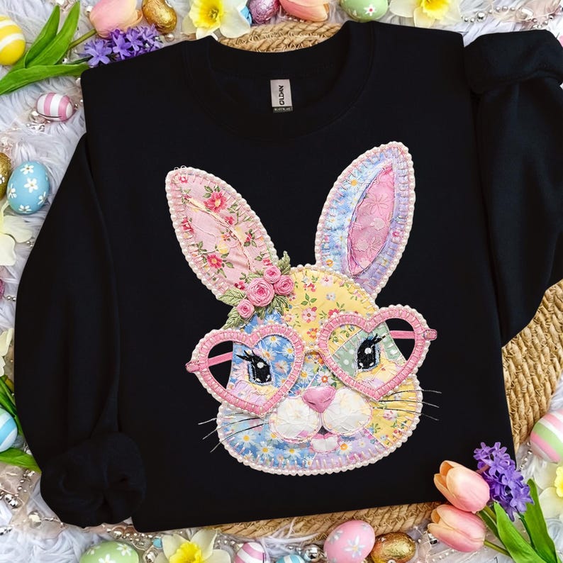 May include: Black sweatshirt featuring a colorful bunny face applique. The bunny has heart-shaped glasses and floral patterned ears. The design includes pink, yellow, blue, and green fabrics. The sweatshirt is surrounded by Easter decorations.